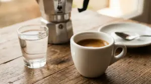 Tazzina di espresso con bicchiere d’acqua su tavolo in legno, con moka sullo sfondo