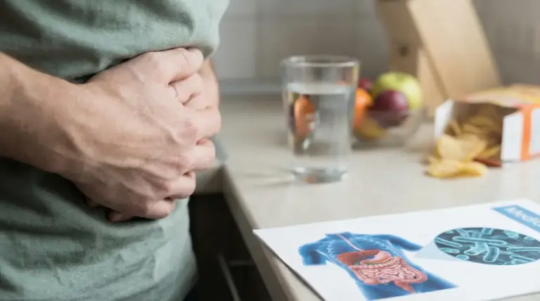 Persona con le mani sull’addome, bicchiere d’acqua e illustrazione dell’apparato digerente sul tavolo