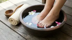 Piedi immersi in una bacinella di acqua fredda con fiori, su pavimento in legno accanto a un asciugamano