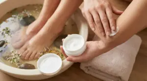 Persona applica crema idratante sul gomito mentre i piedi sono in ammollo in una bacinella con acqua e fiori