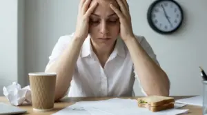Donna stressata con le mani alla testa alla scrivania, tra documenti, caffè e pranzo veloce