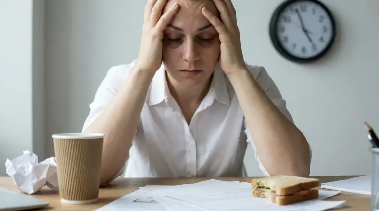 Donna stressata con le mani alla testa alla scrivania, tra documenti, caffè e pranzo veloce