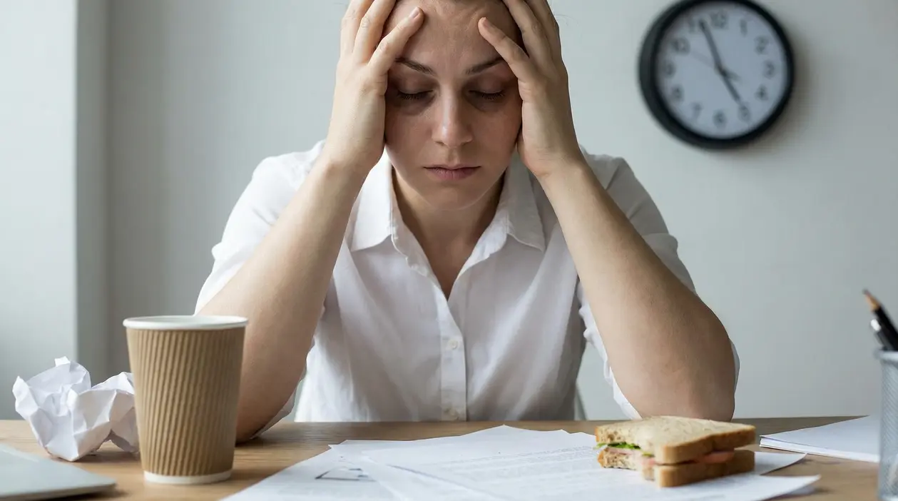 Donna stressata con le mani alla testa alla scrivania, tra documenti, caffè e pranzo veloce